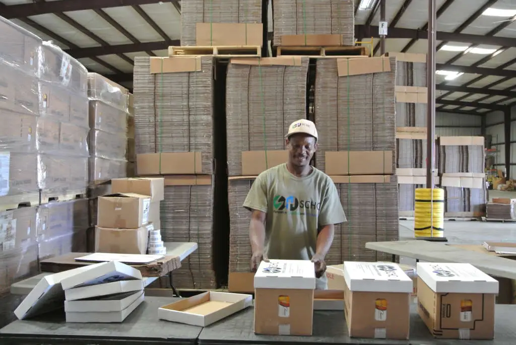 A smiling worker in a cap and logo t-shirt stands among cardboard boxes in a warehouse.