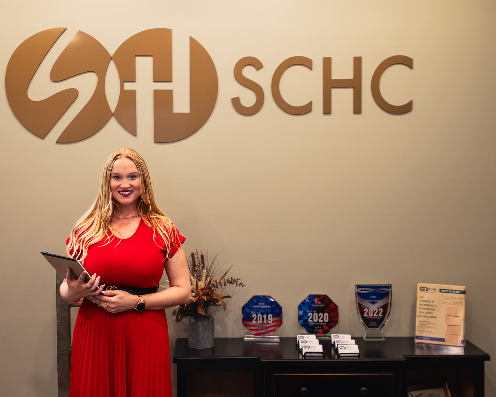 Woman in red dress holding tablet in front of company logo and awards.